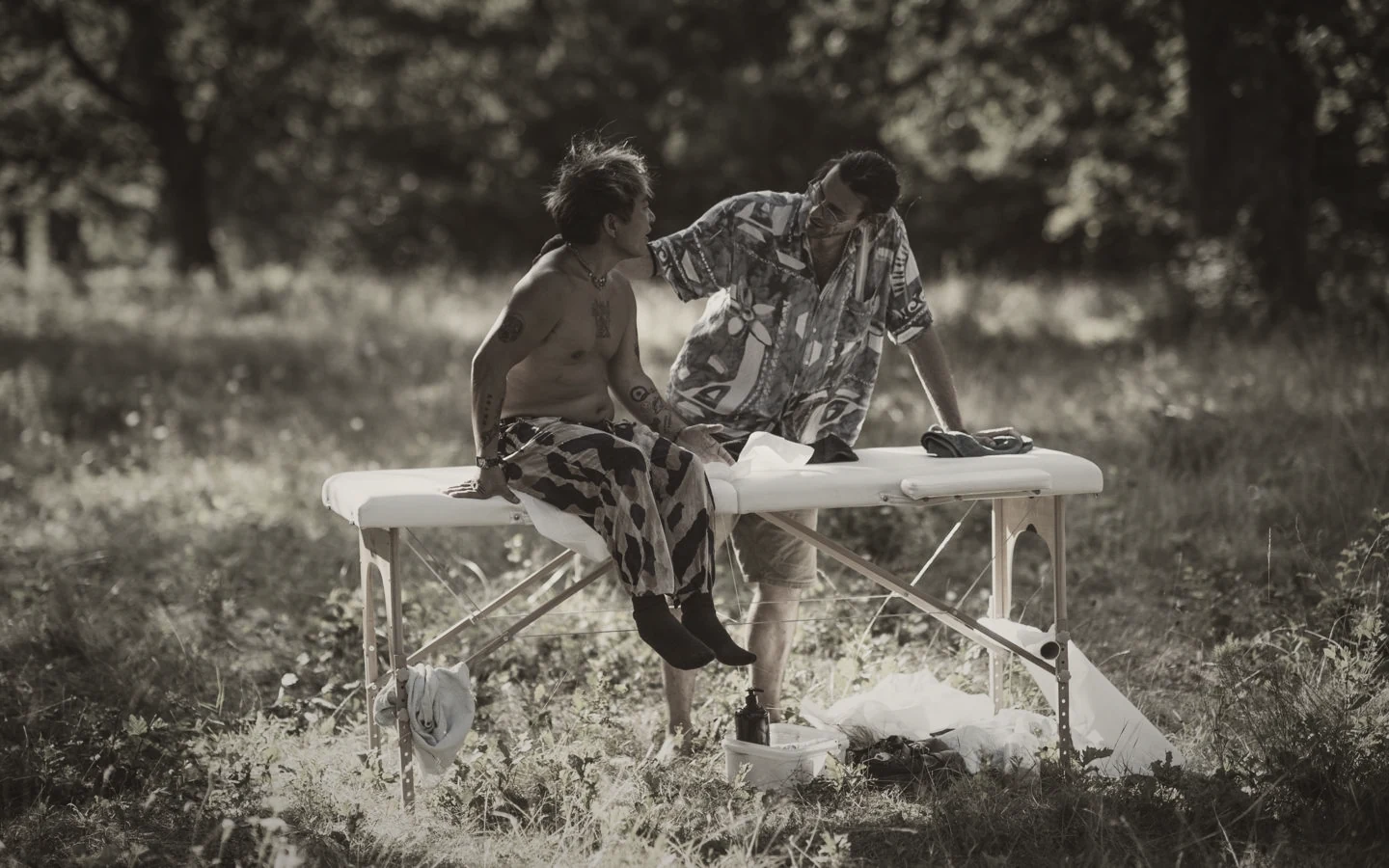 An outdoor massage table set up in a sunlit clearing at a festival, therapist and guest talking beside it.